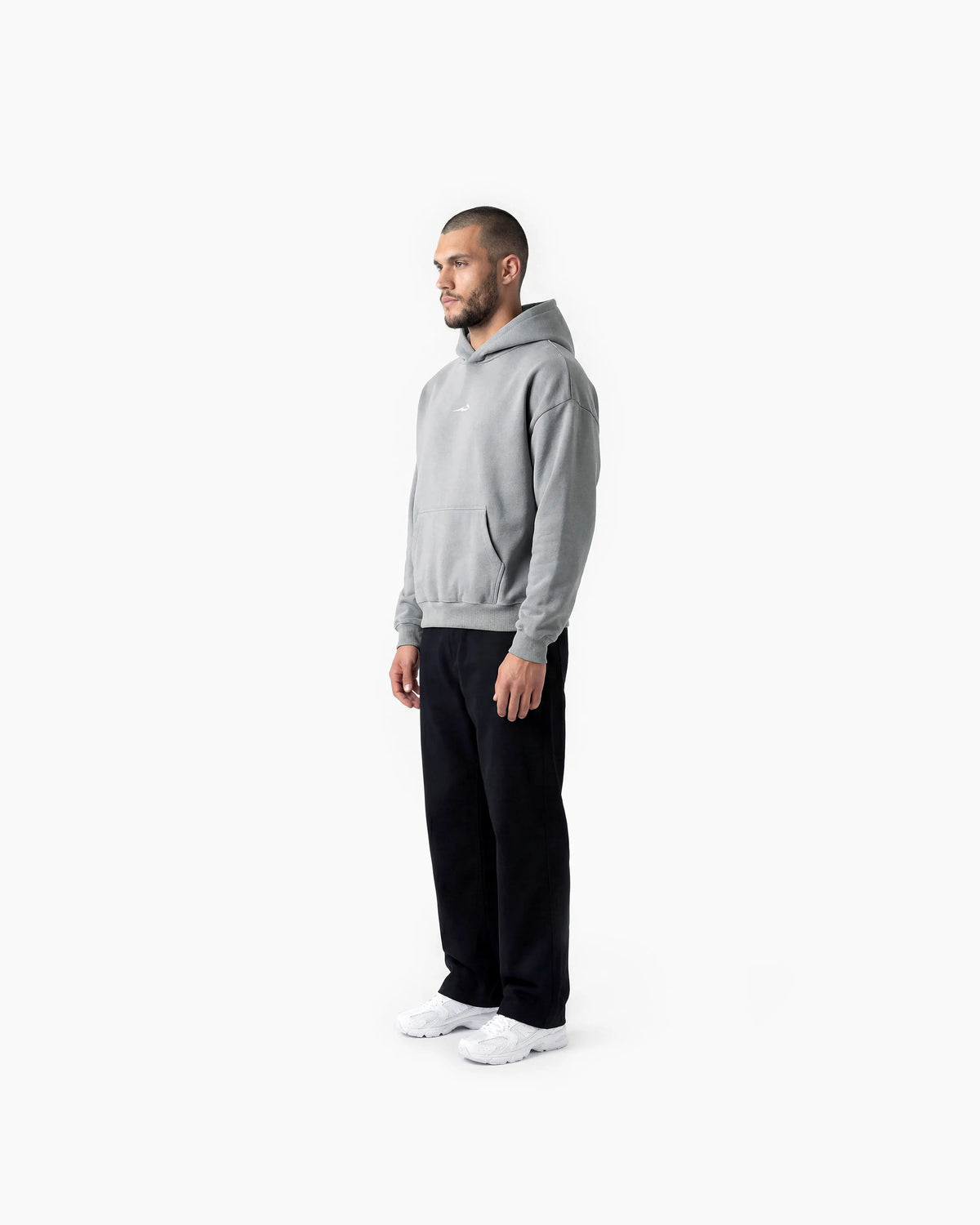 A man in a light grey pullover hoodie, black sweatpants, and white sneakers, shown in profile against a plain white studio background.